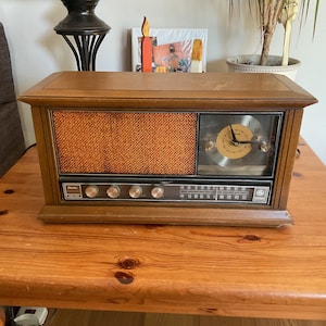 May include: A rectangular wooden radio with a brown textured speaker grille and an integrated analogue clock face. The front panel features a horizontal radio frequency display, five silver control knobs, and the GE logo on the bottom right corner.