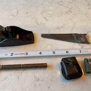 May include: A selection of antique woodworking tools, including a black hand plane, a small saw, a hammer, a leather case, and a metal tool. A ruler shows measurements in inches, equivalent to cm.