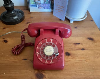 May include: A vintage red rotary telephone featuring a circular dial with numbers and letters, a matching coiled cord, and a handset resting on top. The center of the dial displays a white label with the handwritten text 225-9257 in black ink.