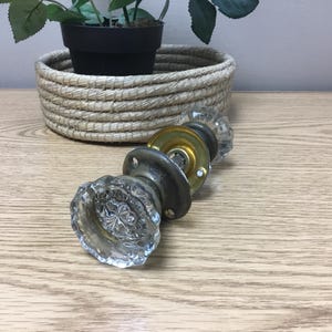 May include: Vintage glass door knobs with brass fittings. The knobs feature a clear, faceted design. The fittings have a tarnished appearance, giving them a vintage aesthetic. A decorative basket with a plant is in the background.