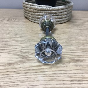 May include: Clear glass door knob with a faceted design. The knob has a brass-coloured base and a metal rod connecting the two knobs. The knobs are lying on a light-coloured wooden surface.