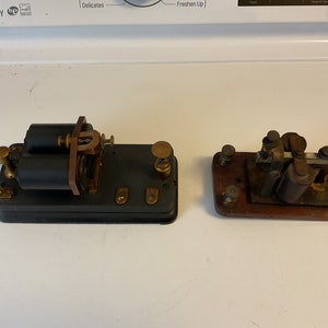 May include: Two antique telegraph keys, one with a black base and two spools, the other with a wooden base and a single spool. Both keys have brass accents.
