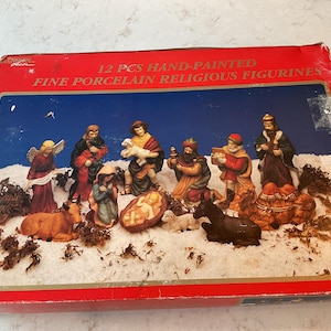 May include: A rectangular red box featuring a 12-piece hand-painted fine porcelain religious figurine nativity set. The box displays an illustration of figures including an angel, Mary, Joseph, the infant Jesus, three kings, a camel, and livestock.