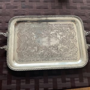 May include: A rectangular silver serving tray with detailed floral and scrollwork engraving. The tray features two curved handles and a rope-style border. The centre of the tray has the text "1950".