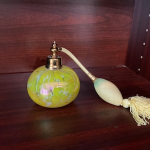May include: A vintage perfume bottle with a yellow and iridescent glass body. It has a gold-coloured metal top and a cream-coloured atomizer with a tassel. The bottle is displayed on a dark wooden surface.
