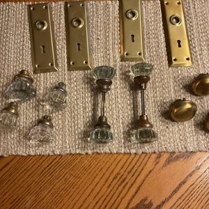 Peut inclure: Un assortiment de quincaillerie de porte ancienne, comprenant des plaques de laiton avec des trous de serrure, des boutons de porte en verre et des boutons de meuble en laiton. L'ensemble est disposé sur une surface beige texturée.