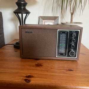 May include: A retro brown radio featuring a speaker grill and a control panel with dials and a tuning display. The radio is placed on a wooden surface. The radio has the brand name "Sony" on the top left corner.
