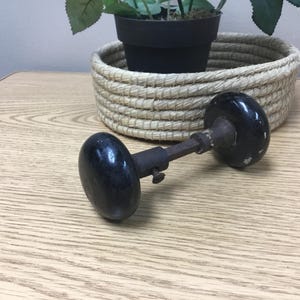 May include: An antique black door knob with a dark, glossy finish. The knob is connected by a metal rod and screw. A woven basket and a potted plant are in the background.