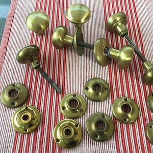 May include: A selection of antique brass door knobs and backplates. The knobs have a rounded design and are in various stages of assembly. The backplates are circular with a central hole. The items are arranged on a red and white striped surface.