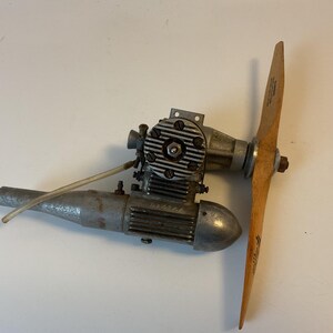 May include: A silver model aircraft engine with a wooden propeller. The engine has a silver exhaust pipe and a silver cylinder head. The propeller is brown and has a white label on it.