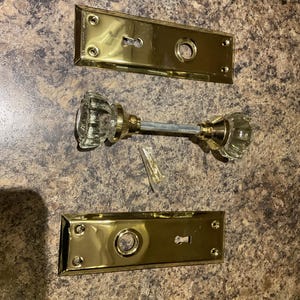 Peut inclure: Un ensemble de quincaillerie de porte vintage comprenant un bouton de porte en verre transparent avec une tige en métal de couleur laiton et deux plaques arrière rectangulaires de couleur laiton. De petites vis en laiton sont également incluses. L'ensemble est sur une surface marron mouchetée.