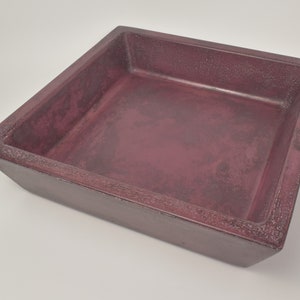 May include: A square, burgundy-colored tray with a textured surface. The tray has a slightly distressed appearance, with visible variations in the color and texture. The sides are angled, and the overall design is simple and functional.