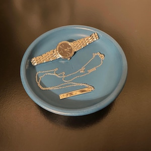 May include: A blue ceramic dish with a silver watch and a silver necklace. The watch has a black face and a silver band. The necklace is a simple chain with a silver bar pendant.
