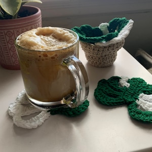 May include: A glass mug filled with a frothy brown beverage, resting on a white crocheted coaster shaped like a four-leaf clover. Two additional crocheted coasters, one green and white and one beige, are also visible.