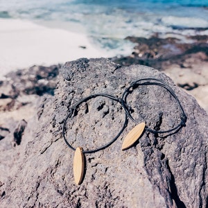 May include: Two black cord bracelets with wooden surfboard pendants rest on a gray rock. The background features a bright blue ocean and a clear sky, suggesting a beach or coastal theme. The bracelets are simple and minimalist.
