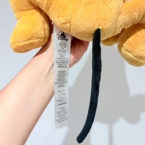 May include: A plush toy in shades of yellow and orange, featuring a long, black tail. The toy has a tag with the Disney logo. The plush appears soft and cuddly, with visible stitching details.