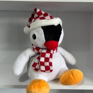 May include: A plush white penguin toy with a black face, red beak, and orange feet. It wears a red and white checkered scarf and a matching hat with a white pom-pom. The toy is sitting on a white shelf.