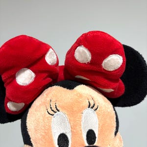 May include: A plush Minnie Mouse doll featuring a red bow with white polka dots. The doll has a peach-colored face, black eyes, and a black nose. The ears are black, and the doll is set against a plain background.