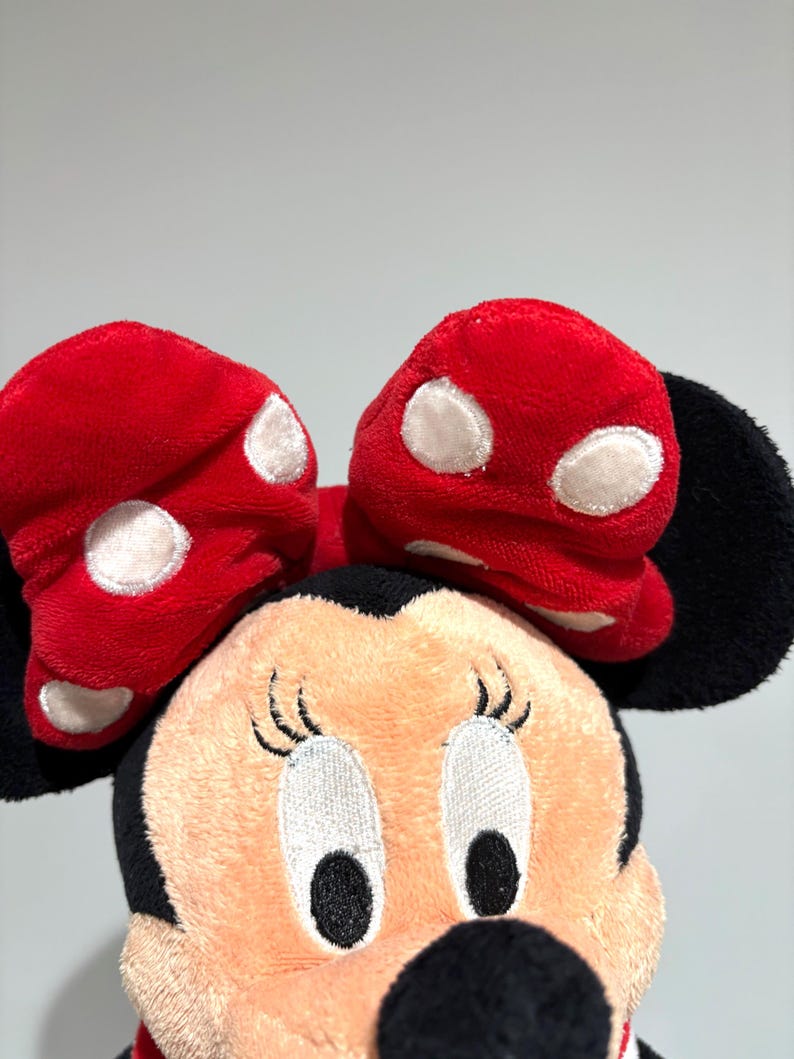 May include: A plush Minnie Mouse doll featuring a red bow with white polka dots. The doll has a peach-colored face, black eyes, and a black nose. The ears are black, and the doll is set against a plain background.