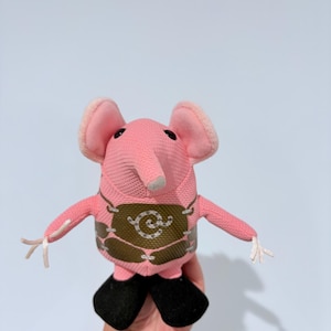 Puede incluir: Un juguete de peluche de elefante rosa con orejas grandes, una trompa gris y pies negros. El elefante lleva un chaleco marrón con un diseño decorativo. El juguete se sostiene en una mano sobre un fondo azul claro.