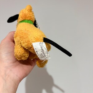 Puede incluir: Un peque&ntilde;o peluche de Pluto con cuerpo amarillo, orejas y cola negras y un collar verde. El juguete tiene una etiqueta que dice "Disneyland Resort Paris".