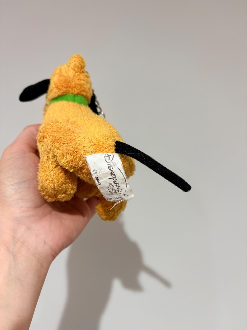Puede incluir: Un peque&ntilde;o peluche de Pluto con cuerpo amarillo, orejas y cola negras y un collar verde. El juguete tiene una etiqueta que dice "Disneyland Resort Paris".