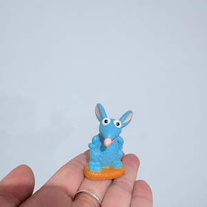 May include: A blue cartoon character figurine with large eyes and a pink tongue, standing on an orange base. The figurine is held in a person's hand, with silver rings on the fingers.