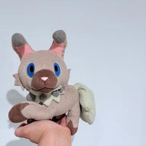 Puede incluir: Un gato de peluche con cuerpo marrón claro, orejas grises y ojos azules. El gato tiene una cola verde claro y un collar verde claro y gris. El juguete está en la mano de una persona.