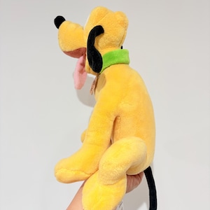 May include: A plush toy of Pluto, a yellow dog with black ears and tail, a green collar, and a pink tongue. The stuffed animal is a Disney character.