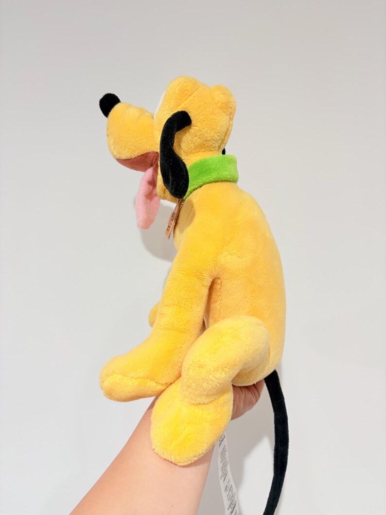 May include: A plush toy of Pluto, a yellow dog with black ears and tail, a green collar, and a pink tongue. The stuffed animal is a Disney character.