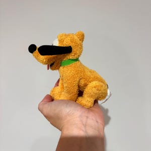 Puede incluir: Un peluche de un perro amarillo con nariz y orejas negras, un collar verde y una lengua roja. El perro est&aacute; sentado y tiene una mancha blanca en la cabeza. El juguete es un personaje de dibujos animados.