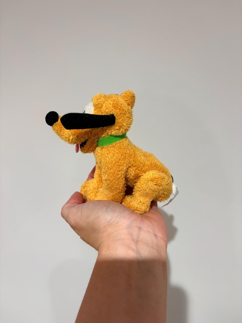 Puede incluir: Un peluche de un perro amarillo con nariz y orejas negras, un collar verde y una lengua roja. El perro est&aacute; sentado y tiene una mancha blanca en la cabeza. El juguete es un personaje de dibujos animados.