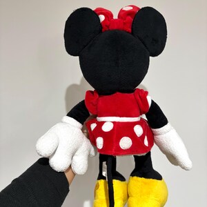 May include: A plush Minnie Mouse doll, viewed from the back, features a red dress with white polka dots, a matching bow, and yellow shoes. The doll has black ears, arms, and legs, with white gloves. The doll is held against a plain background.