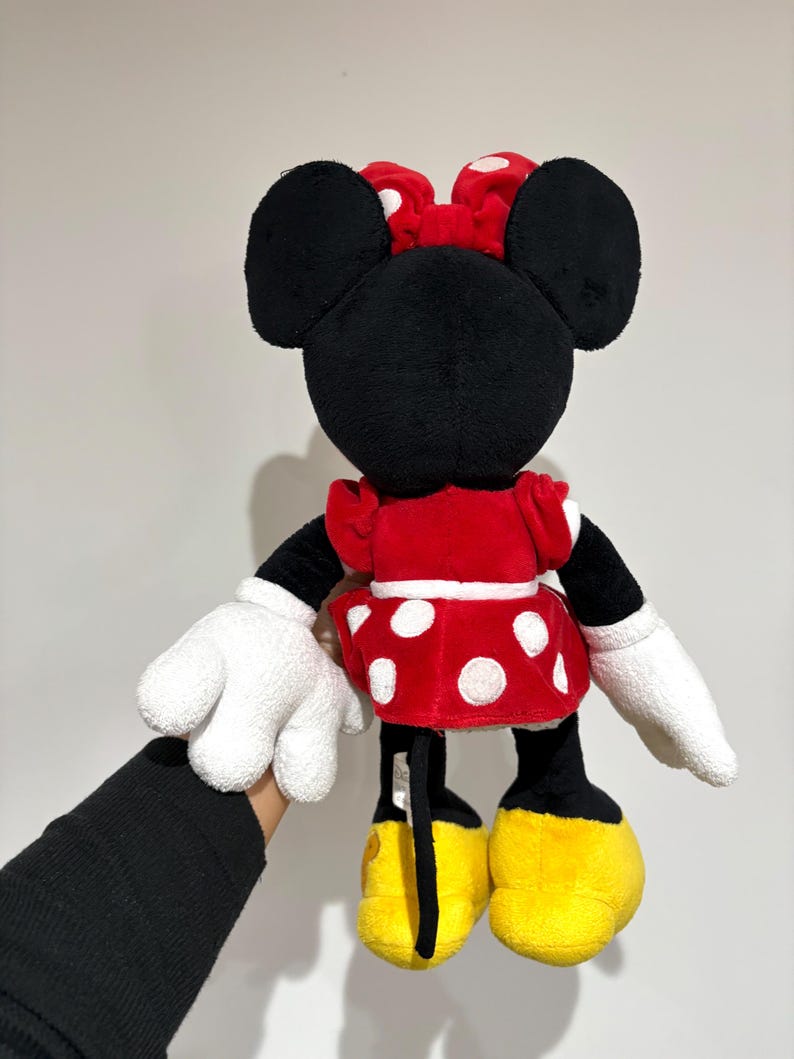 May include: A plush Minnie Mouse doll, viewed from the back, features a red dress with white polka dots, a matching bow, and yellow shoes. The doll has black ears, arms, and legs, with white gloves. The doll is held against a plain background.