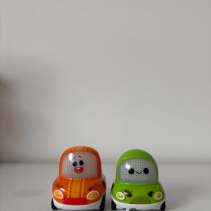 May include: Two toy cars, one orange with yellow stripes and the other green, sit side-by-side. Each car has a smiling face on the front and black wheels. The cars are designed with rounded shapes and a playful aesthetic.