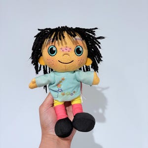 Puede incluir: Una muñeca de peluche con una cara redonda y amarilla, cabello negro de hilo y ojos azules. La muñeca lleva una camisa azul claro con diseños de estrellas, pantalones cortos amarillos, calcetines rosas y zapatos negros. La muñeca está siendo sostenida.