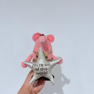 May include: A pink plush toy in the shape of a mouse holding a silver star. The star has the text "you're out of this world" printed on it. The mouse has large pink ears and a brown nose.