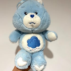 Könnte beinhalten: Hellblauer Plüschbär mit weißem Bauch und blauem Wolkenmuster. Der Bär hat eine herzförmige Nase, weiße Akzente und einen traurigen Gesichtsausdruck. Ein Vintage-Spielzeug.