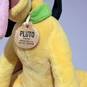 May include: A plush yellow Pluto dog toy with a green collar and a tag that reads "PLUTO" and "if found, please return to MICKEY MOUSE." The toy has a pink tongue, black ears, and a brown nose.