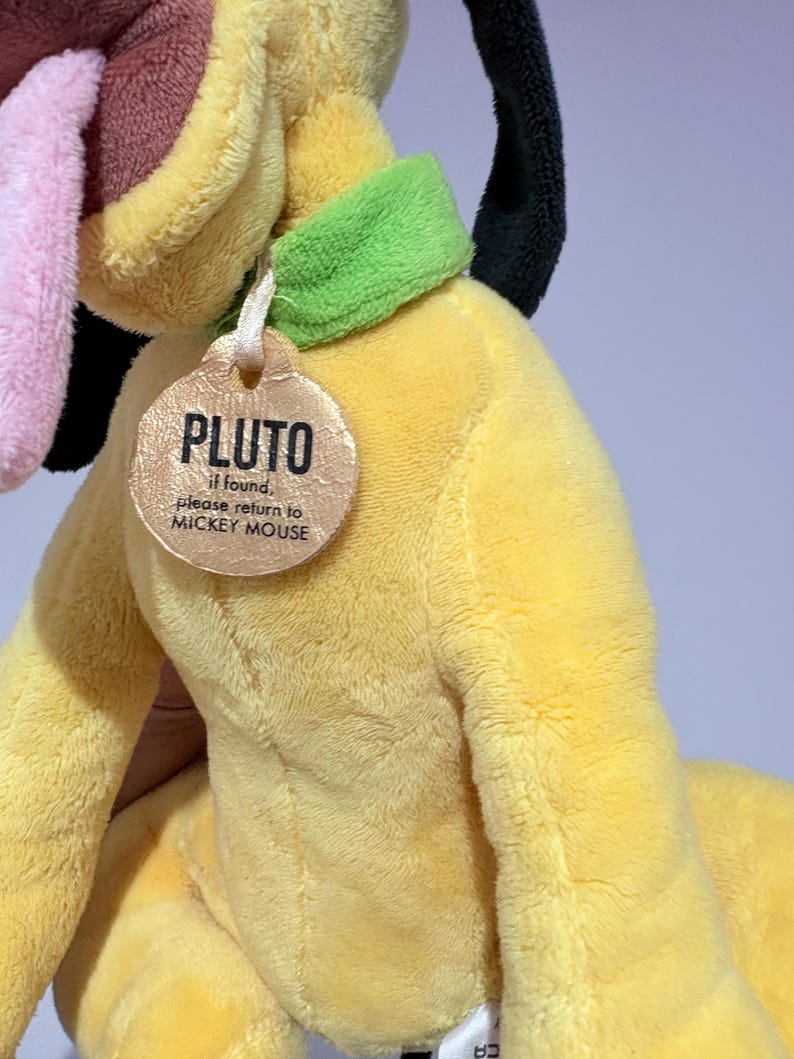 May include: A plush yellow Pluto dog toy with a green collar and a tag that reads "PLUTO" and "if found, please return to MICKEY MOUSE." The toy has a pink tongue, black ears, and a brown nose.