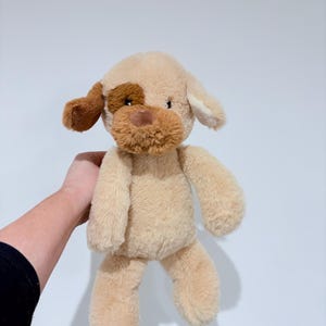 Peut inclure: Une peluche en forme de chien, de couleur beige et marron. Le chien a des oreilles tombantes, une tache marron sur un œil et un museau marron. La peluche est douce et moelleuse.