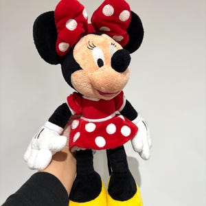 May include: A Minnie Mouse plush doll. The doll has a red dress with white polka dots, a matching bow, and yellow shoes. The doll's face is beige with black eyes and a black nose. The doll is being held by a person.