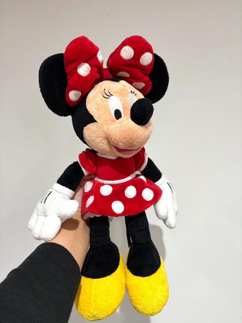 May include: A Minnie Mouse plush doll. The doll has a red dress with white polka dots, a matching bow, and yellow shoes. The doll's face is beige with black eyes and a black nose. The doll is being held by a person.