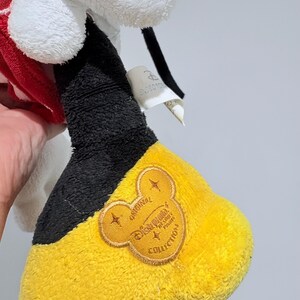 May include: A plush toy featuring a white head, red and white patterned accents, and a black and yellow foot. The foot has a black cuff and a yellow sole with an embroidered Disney Parks Collection logo.