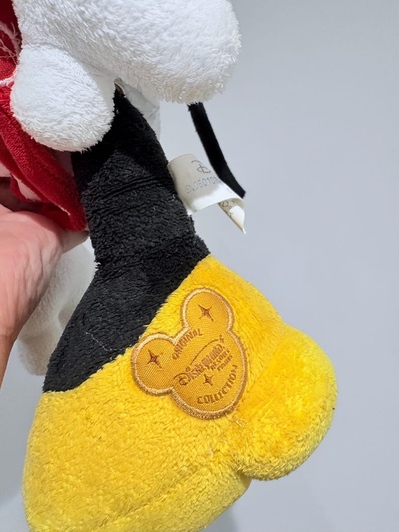 May include: A plush toy featuring a white head, red and white patterned accents, and a black and yellow foot. The foot has a black cuff and a yellow sole with an embroidered Disney Parks Collection logo.