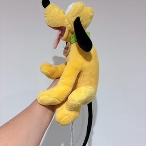 May include: A plush yellow Pluto dog toy with black ears and tail, a pink tongue, and a green collar. The toy has a tag attached to the collar. The dog is in a sitting position.