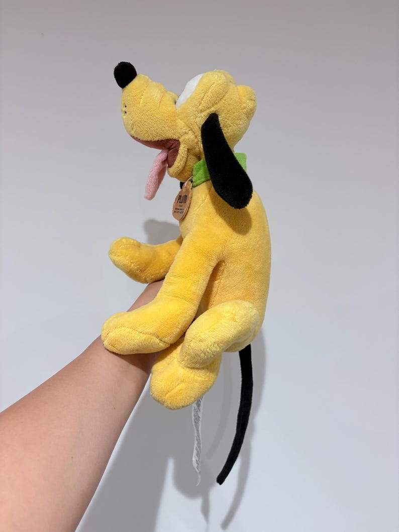 May include: A plush yellow Pluto dog toy with black ears and tail, a pink tongue, and a green collar. The toy has a tag attached to the collar. The dog is in a sitting position.
