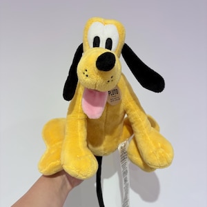 May include: A plush Pluto toy, featuring a yellow body, black ears, and a black nose. The toy has a pink tongue sticking out and a tag that reads "Pluto". The toy is a classic Disney character.