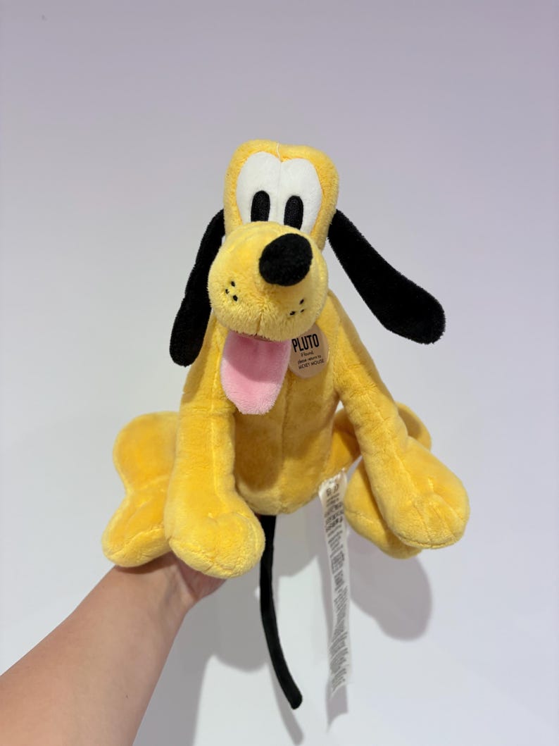 May include: A plush Pluto toy, featuring a yellow body, black ears, and a black nose. The toy has a pink tongue sticking out and a tag that reads "Pluto". The toy is a classic Disney character.