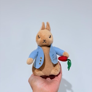 Puede incluir: Un peluche de un conejo beige con una chaqueta azul claro y una zanahoria roja con hojas verdes. El conejo tiene una expresión seria. El juguete se sostiene en una mano sobre un fondo blanco.